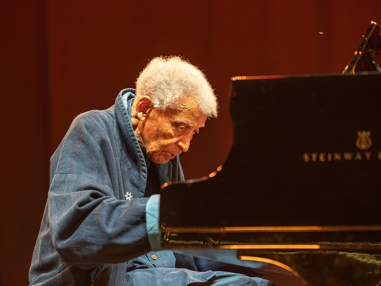 Abdullah Ibrahim playing a grand piano at Rudolstadt Festival in July 2025, retaining the complete warm-lit photograph and instrument.