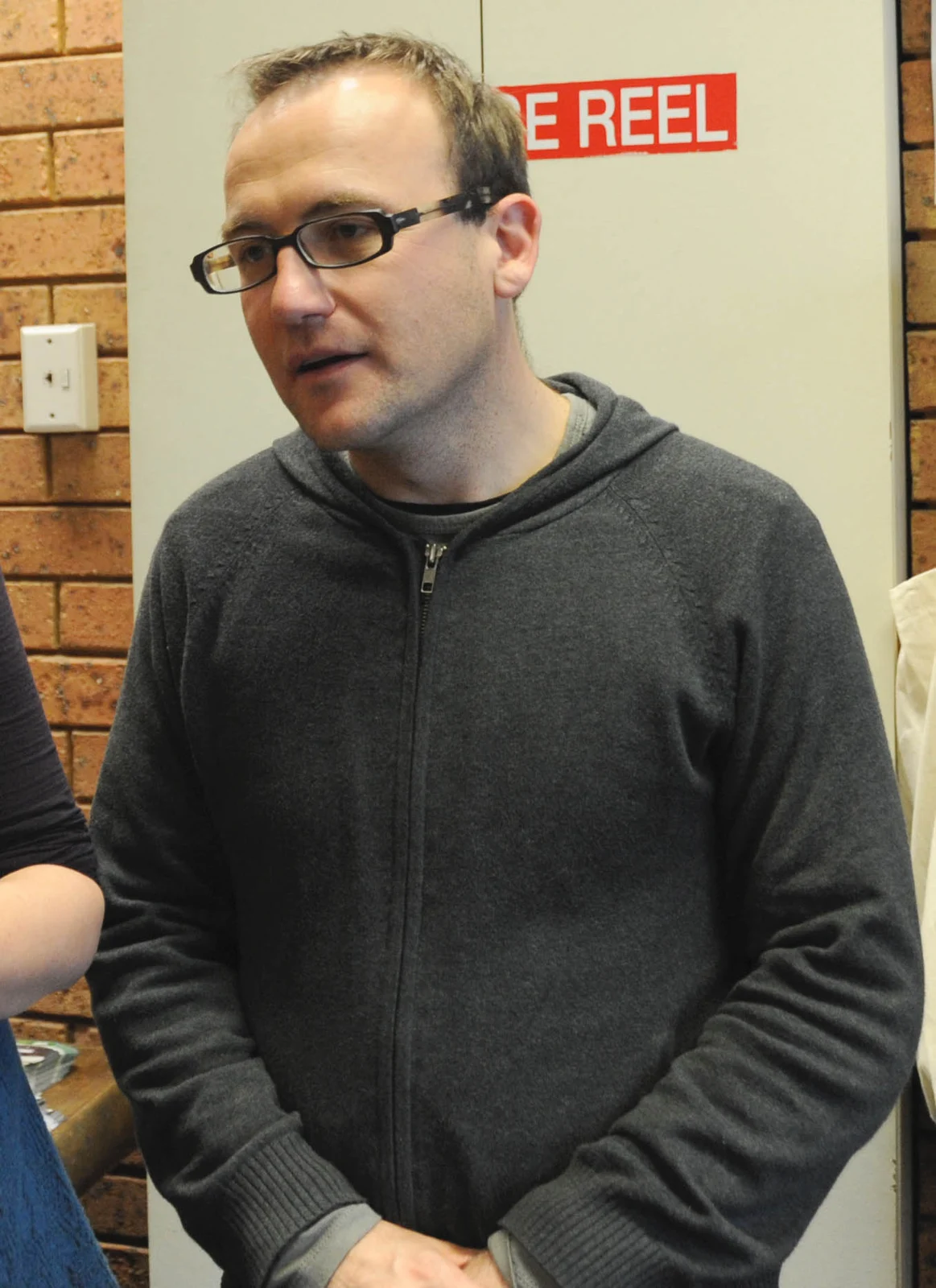 Adam Bandt wearing glasses and a grey zip-up jacket in his campaign office in June 2010, with parts of other people retained at the frame edges.