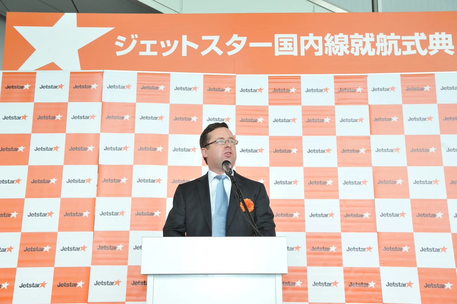 Complete Jetstar photograph of Alan Joyce speaking at a podium at Narita in 2012, retaining the full orange-and-white launch banner and stage frame.