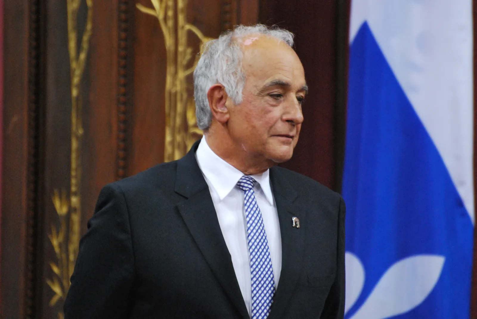 Aldo Bensadoun in a dark suit and patterned tie at a 2013 Québec investiture, with wood panelling and the Québec flag retained in Simon Villeneuve’s full photograph.