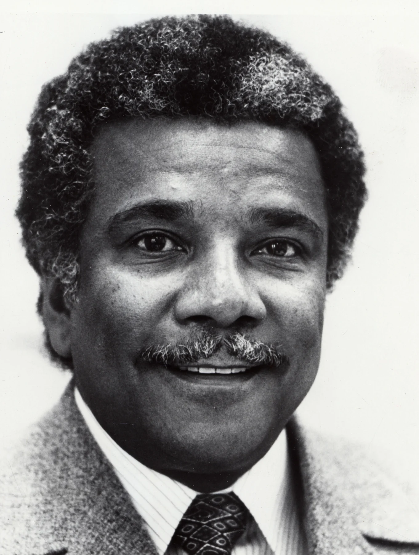 Ali Mazrui in an undated black-and-white archival portrait, wearing a jacket and patterned tie; complete supplied frame.