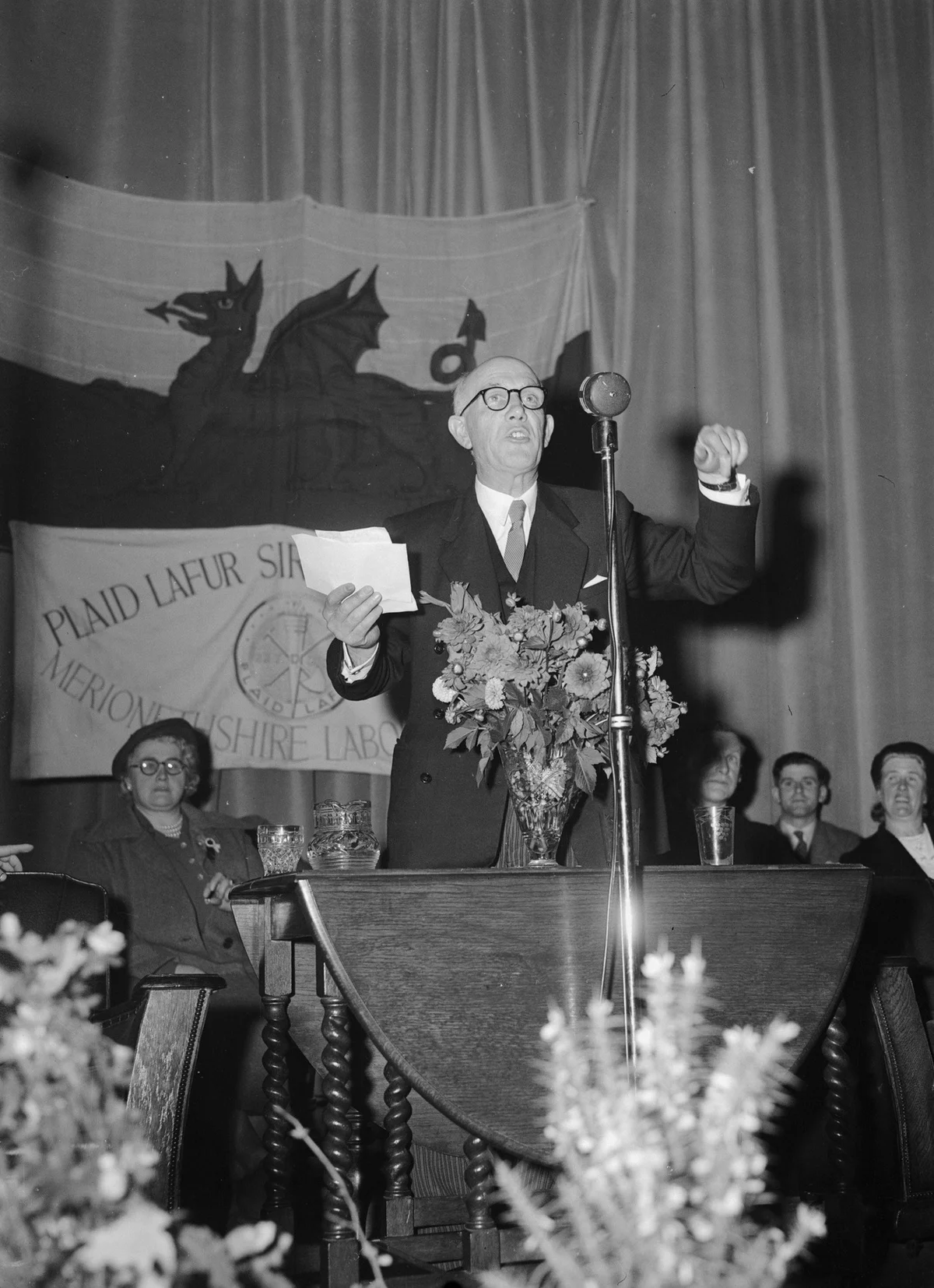 Aneurin Bevan speaking at a Labour meeting in Corwen on 3 October 1952, with the Welsh flag, other attendees and complete supplied photographic frame retained.