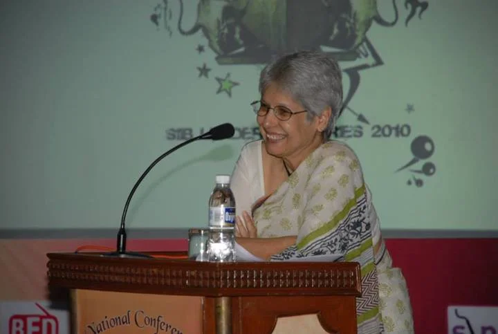 Anu Aga speaking at a conference lectern, with microphones, water bottles and the complete backdrop bearing 2010; exposure date unresolved.
