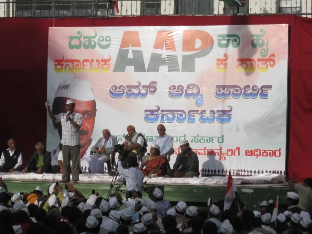 Arvind Kejriwal stands at left holding a microphone at an AAP event in Bangalore in July 2013, with colleagues seated and supporters gathered around the stage.
