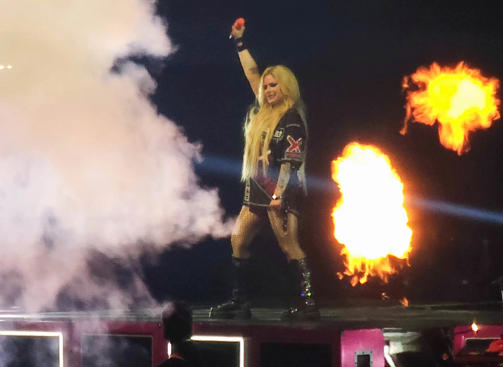 Avril Lavigne raising a microphone on the Budweiser Stage amid smoke and pyrotechnics in August 2024