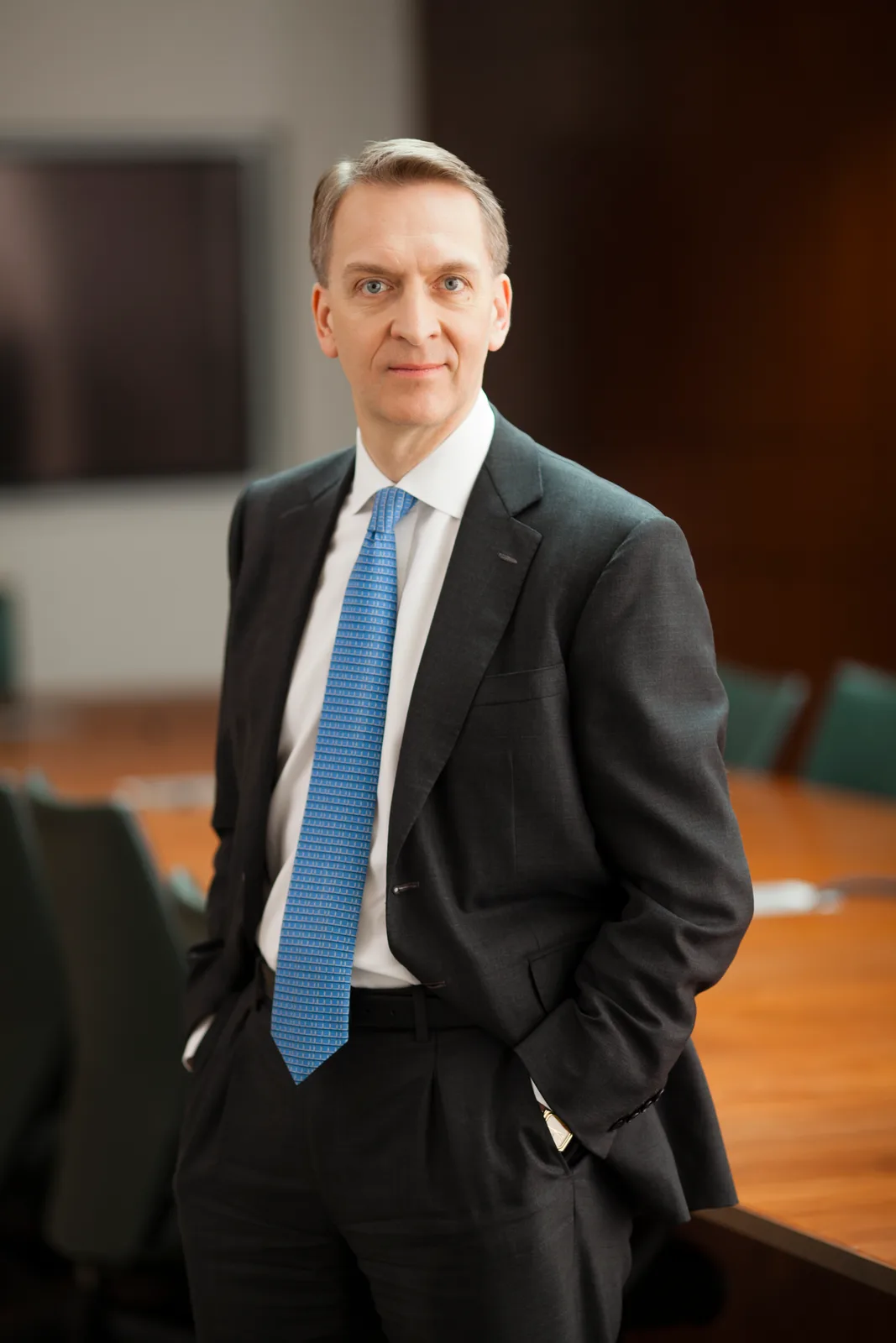 Bruce Flatt stands in a dark suit and blue tie in an office, with both hands in his pockets; Todd France’s complete supplied portrait retains the surrounding room.