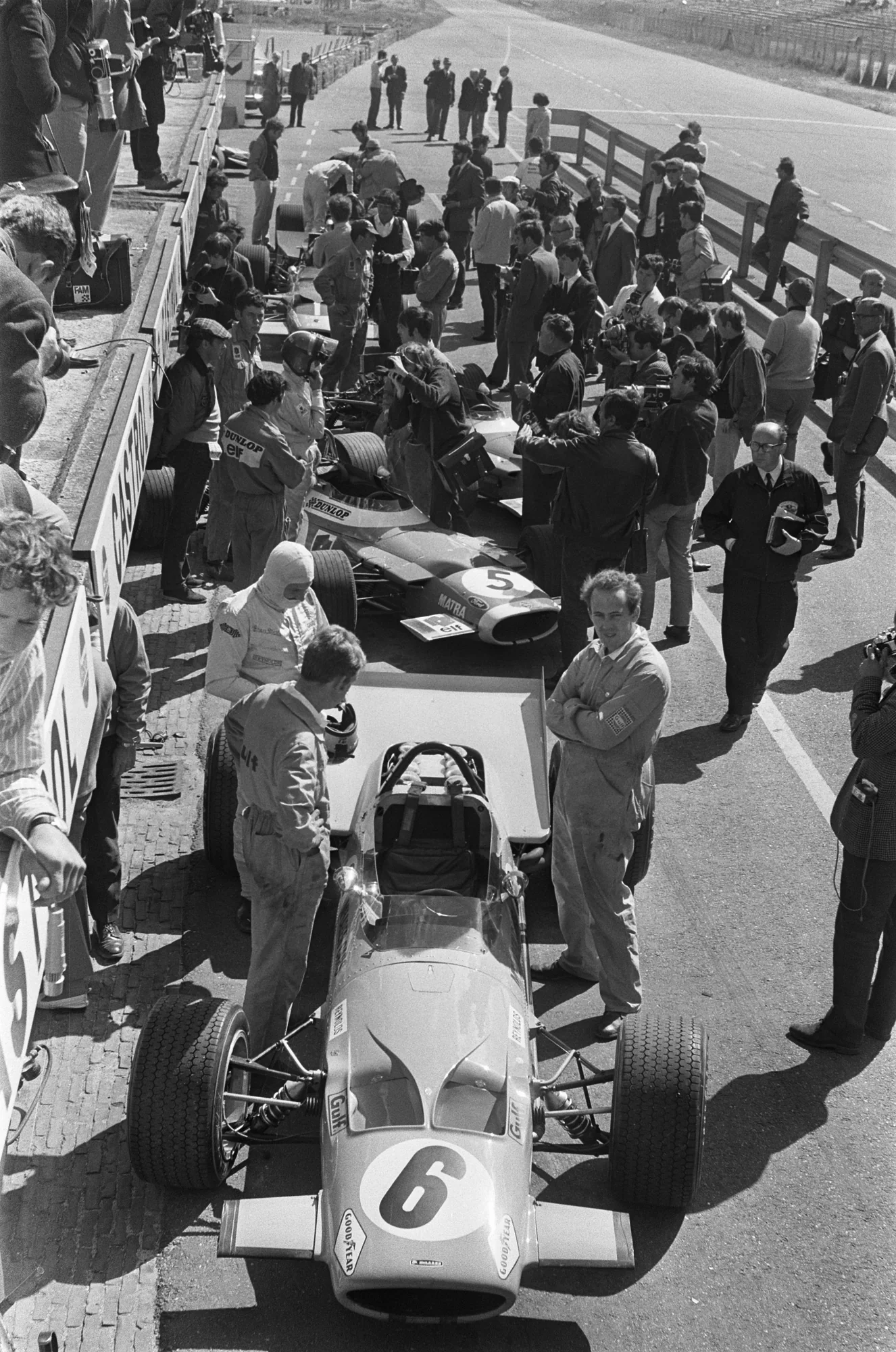 Complete pit-lane photograph at Zandvoort on 19 June 1969, with Bruce McLaren in a white balaclava beside his number 6 car; other people and cars remain visible.