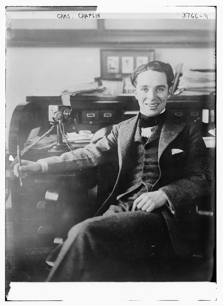 Charlie Chaplin seated smiling at a desk in a Bain photograph catalogued 1915, with telephone, plate border, handwritten name and negative number preserved
