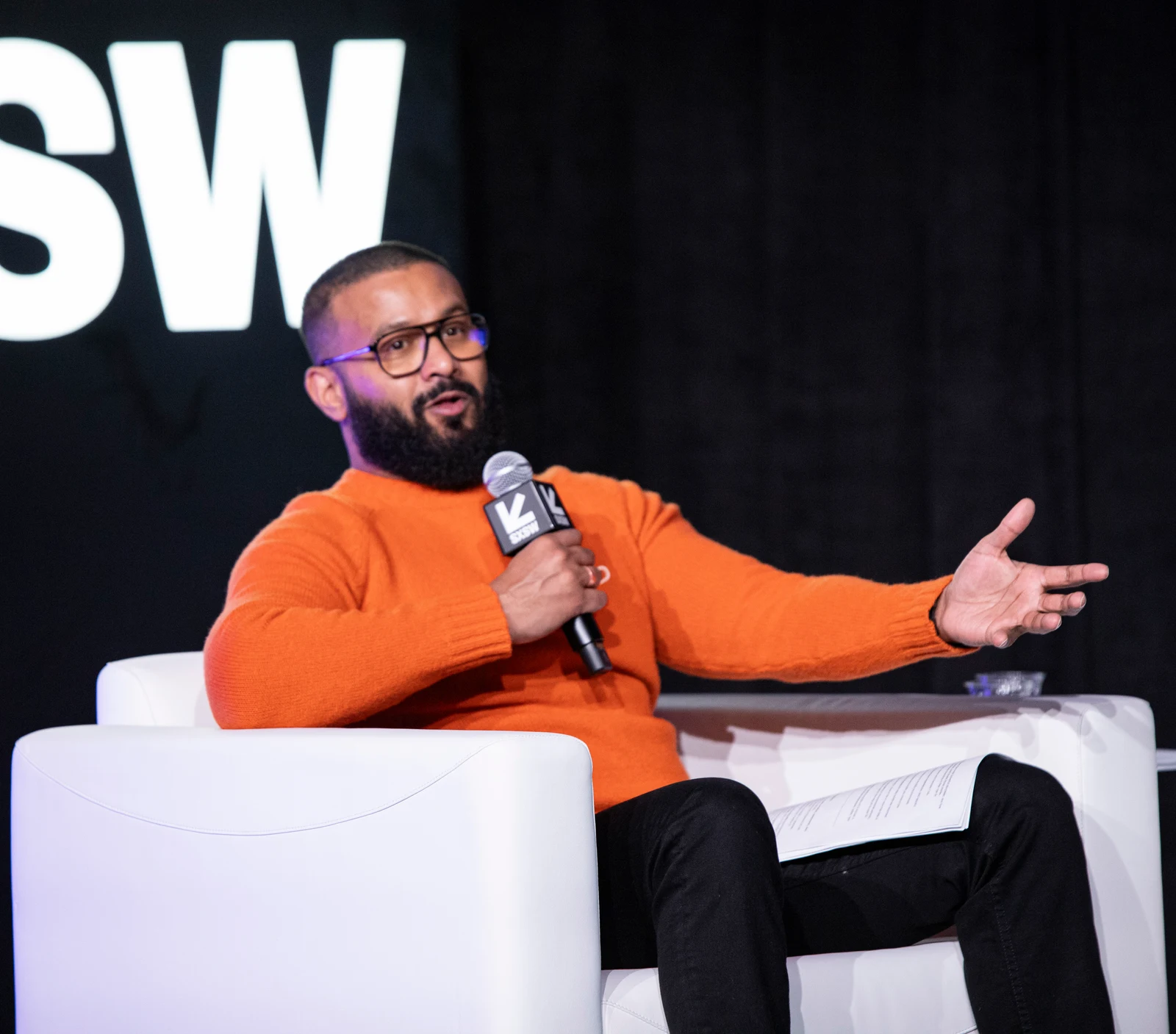 Dax Dasilva seated in an orange sweater and glasses, speaking into a microphone at SXSW in 2025, with his outstretched hand and chair retained.