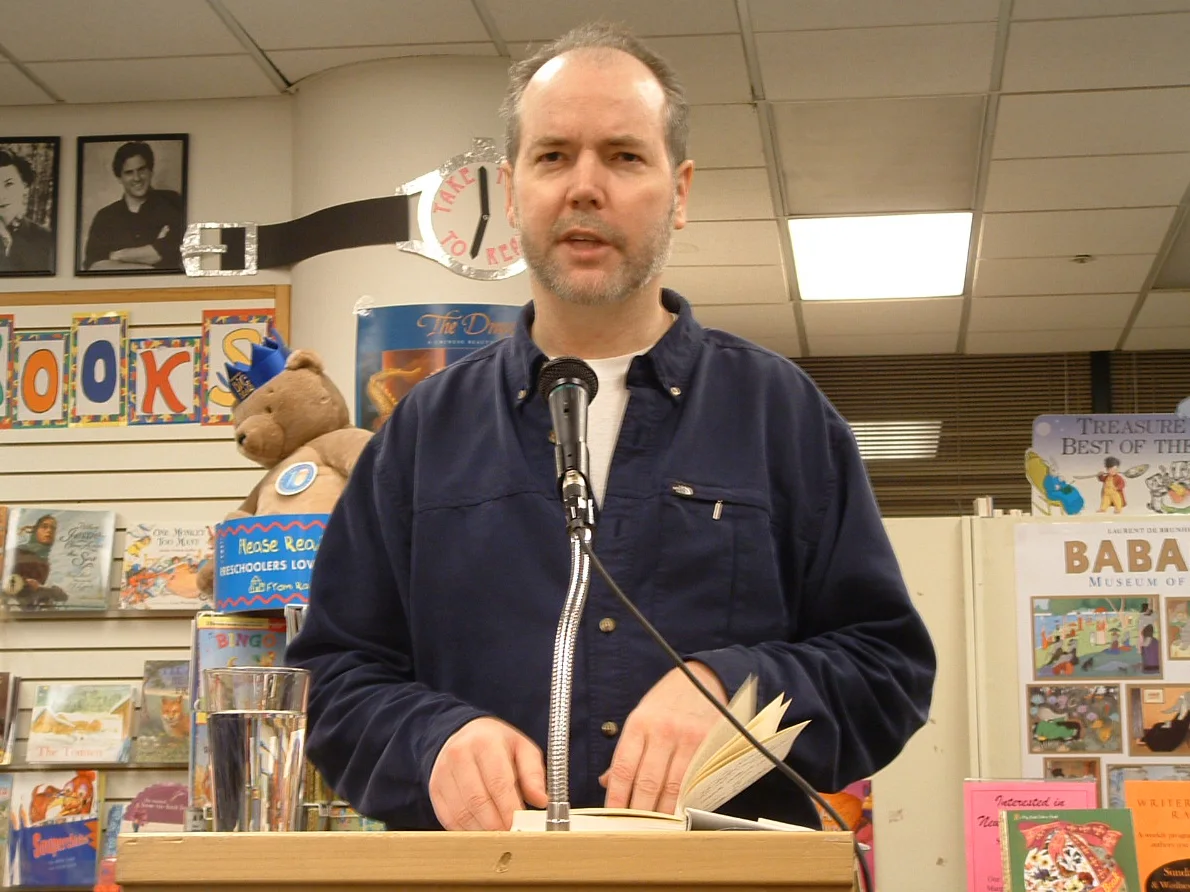 Douglas Coupland in a navy shirt holding an open book behind a microphone at a San Francisco bookstore reading in January 2005