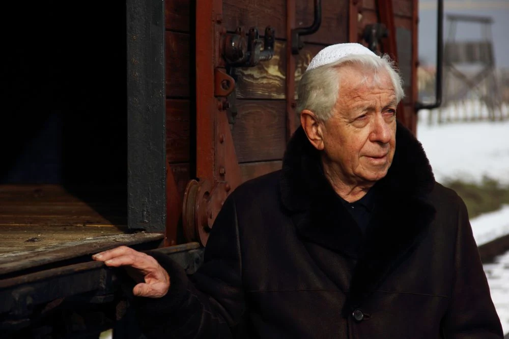 Frank Lowy in a dark coat and white head covering beside a wooden railway wagon at the 2013 March of the Living, with the complete supplied frame retained.