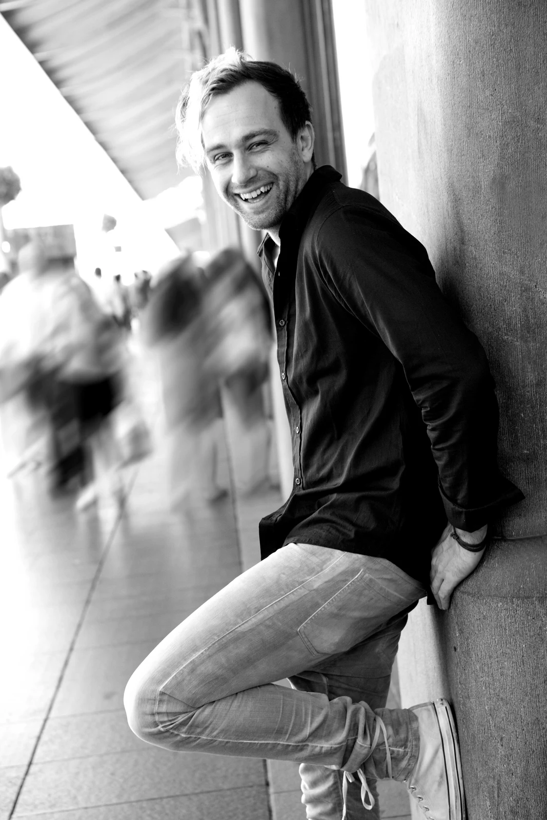 Full black-and-white 2015 Sydney portrait of Fred Schebesta leaning beside a wall, with his shoes, the pavement and background pedestrians retained.