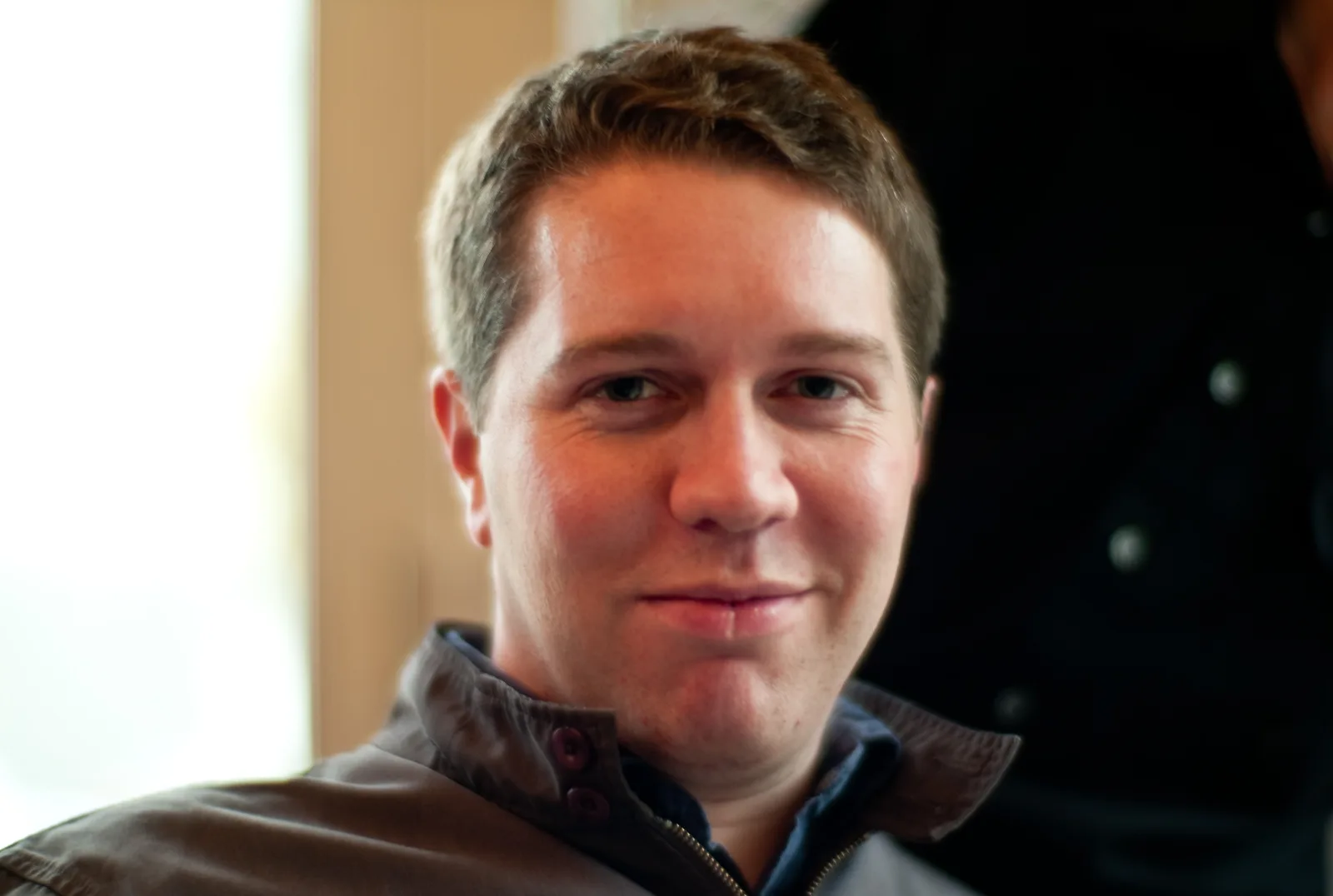 Garrett Camp looks towards the camera in a brown jacket, with a bright window at left, in Joi Ito’s complete 2009 photograph.