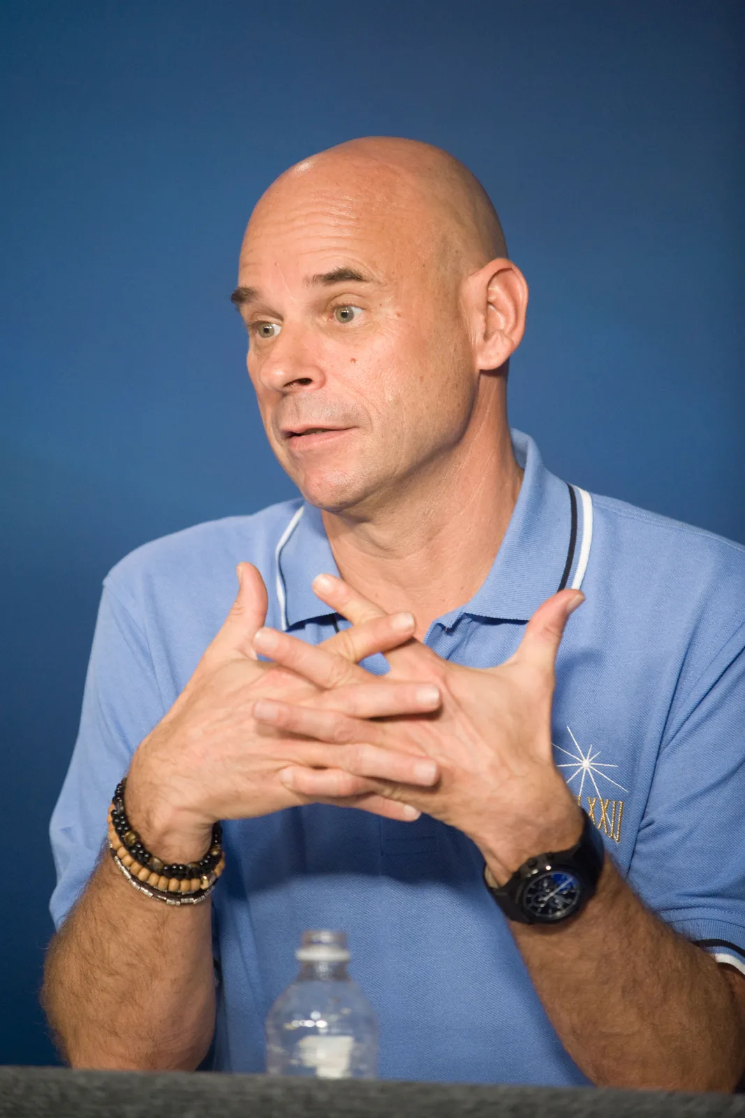 Guy Laliberté, wearing a blue polo shirt, speaks with his hands clasped at a July 2009 Johnson Space Center press conference; the full supplied frame includes the table and water bottle.