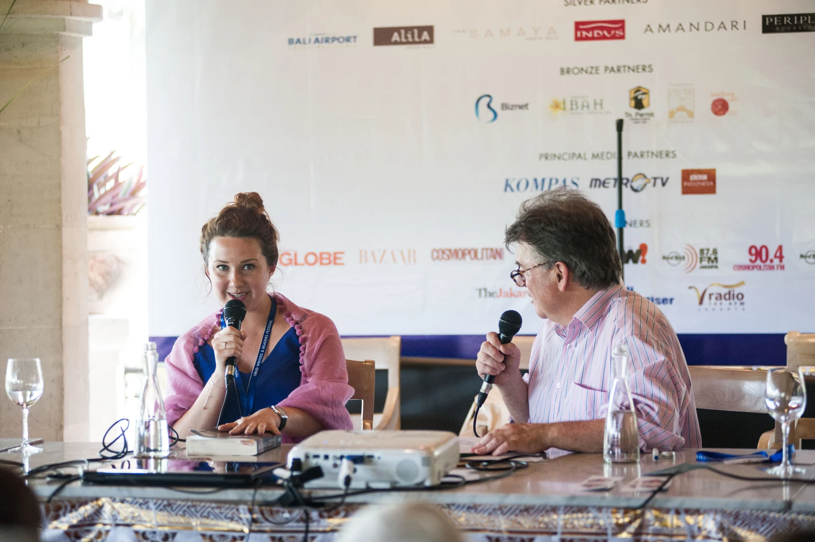 Hannah Kent at left holding a microphone beside another speaker at the 2016 Ubud festival; the full stage, table and backdrop are retained.