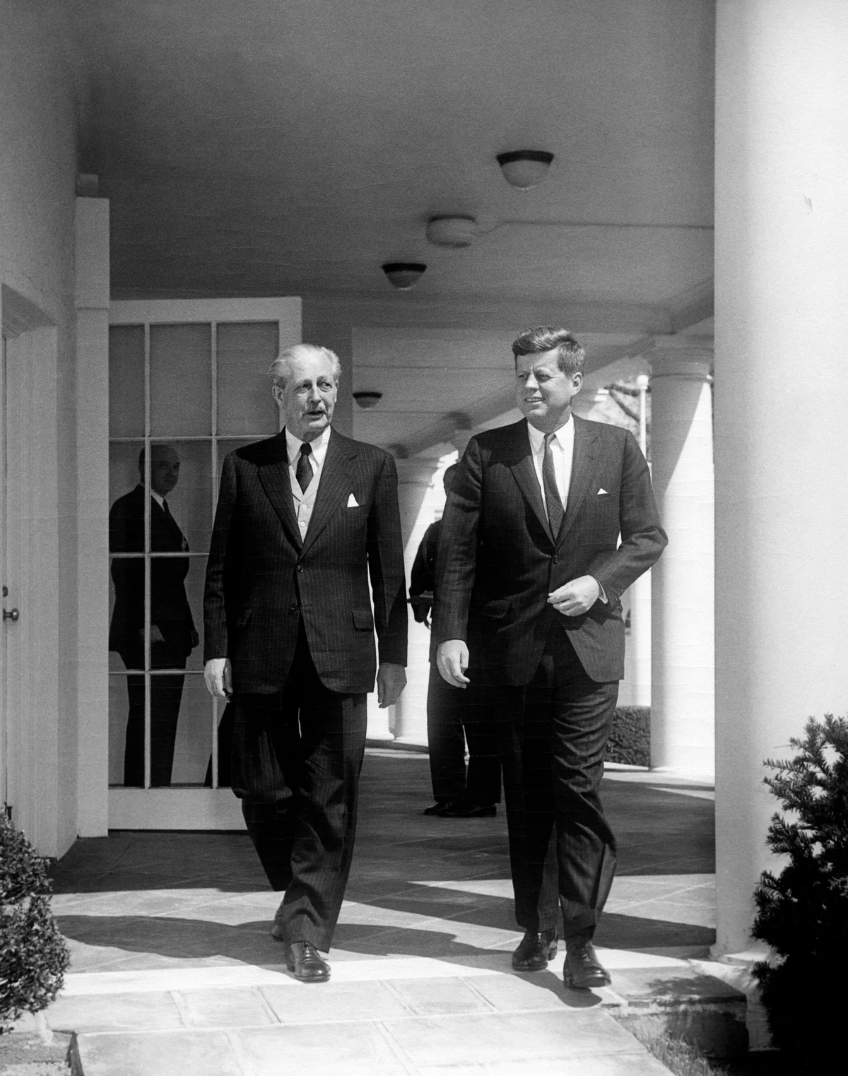Harold Macmillan at left walking with John F. Kennedy in the White House colonnade on 5 April 1961, with other participants visible behind them.