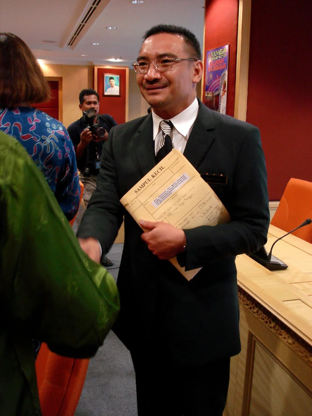Hishammuddin Hussein in a dark suit and glasses, holding a booklet while greeting people at a May 2006 event.