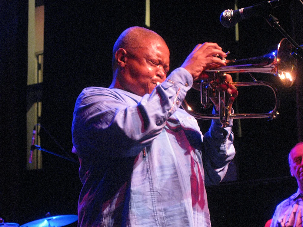 Hugh Masekela playing a horn on stage in April 2009, retaining the full photograph including microphone, drums and another musician at right.