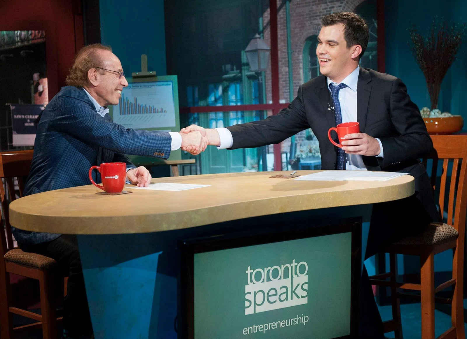 Isadore Sharp, left, shakes hands with Evan Kosiner across a television studio desk in the complete 2013 two-person photograph.