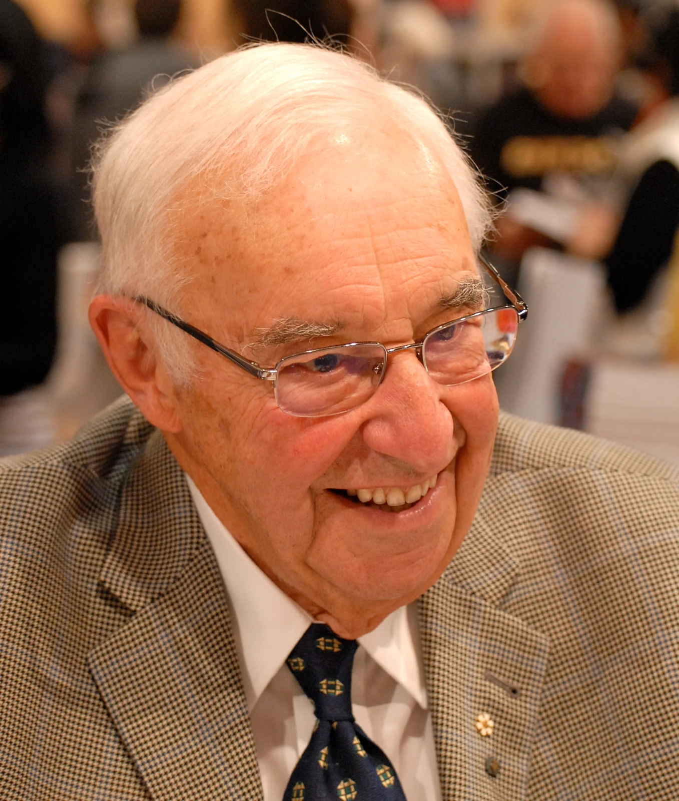Jean Coutu smiling in glasses, a checked jacket and patterned tie at the 2010 Montréal book fair in Asclepias’s full original photograph.