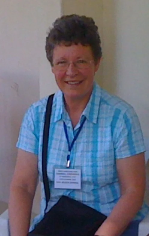 Jocelyn Bell Burnell in a blue checked shirt, wearing a conference badge and holding a dark folder.