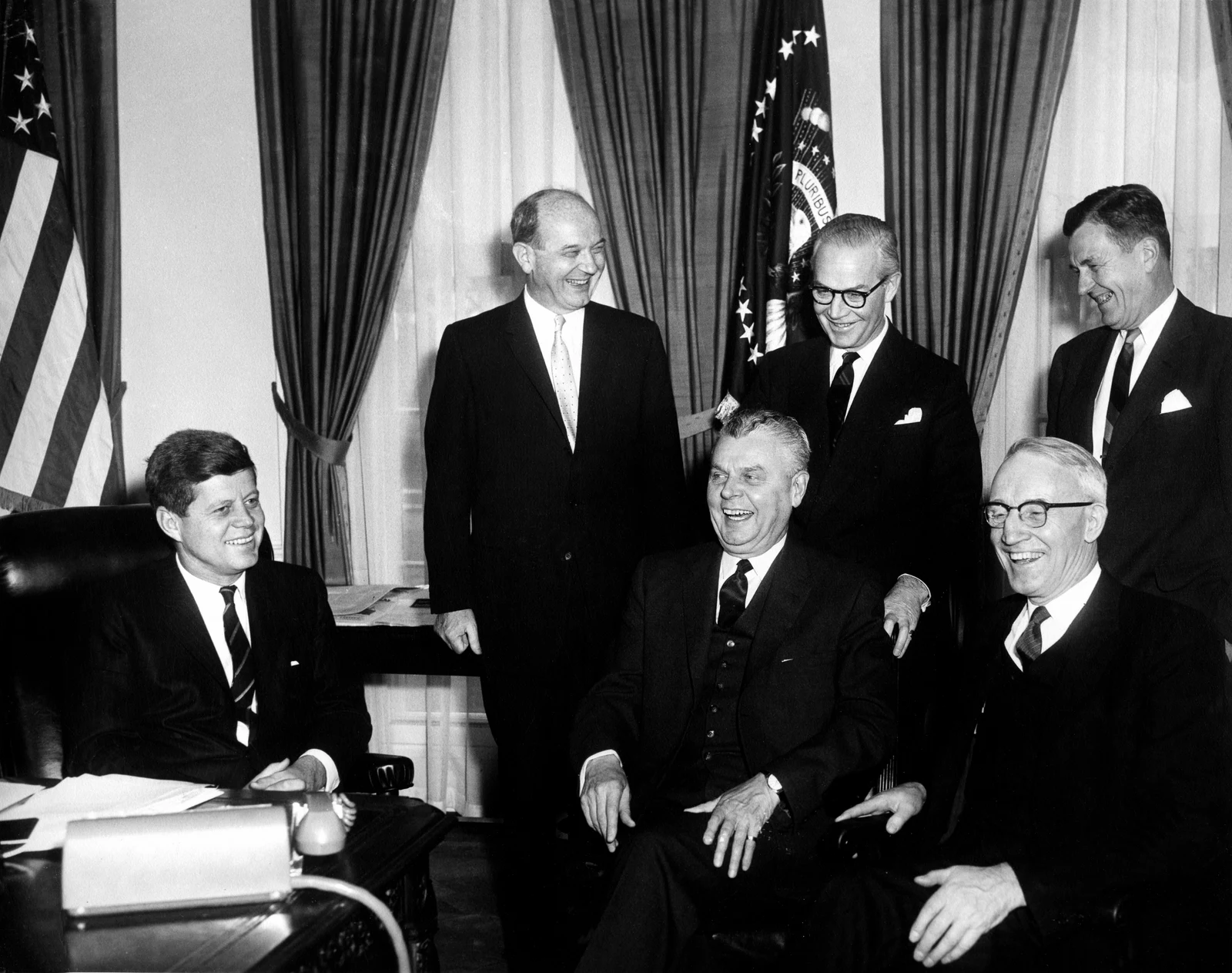 John Diefenbaker seated in the centre beside John F. Kennedy at left and Howard Green at right, with three officials standing behind them in February 1961; the full six-person frame is retained.