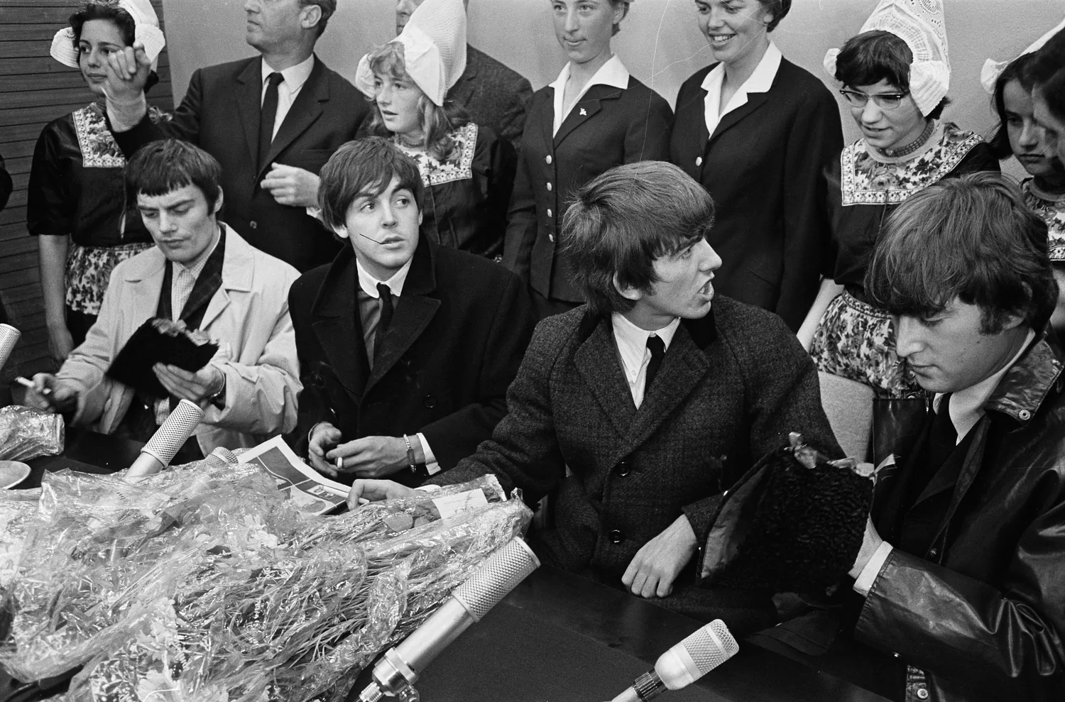 Complete 1964 Schiphol press photograph with John Lennon at the far right, beside George Harrison, Paul McCartney and temporary drummer Jimmie Nicol, retaining the gathered people and microphones