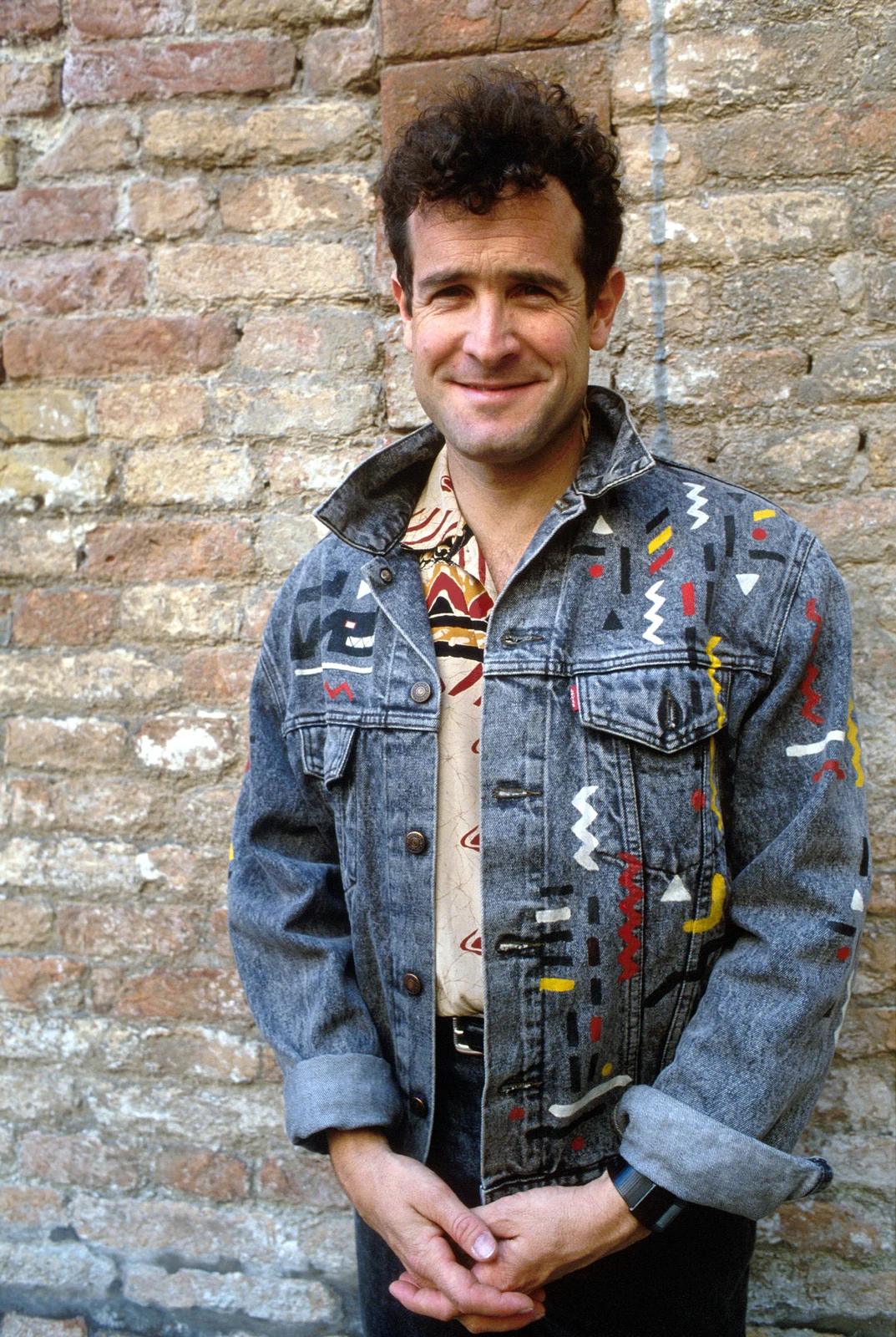 Johnny Clegg standing before a stone wall in the complete 1992 portrait, retaining his jacket and clasped hands.