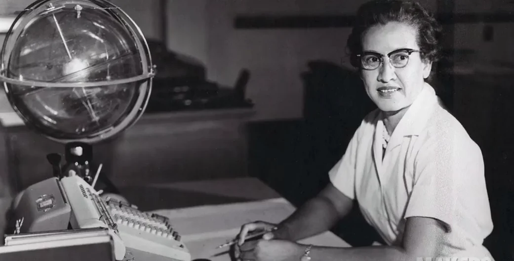 Katherine Johnson seated at her desk with papers, a calculating machine and a celestial training device.