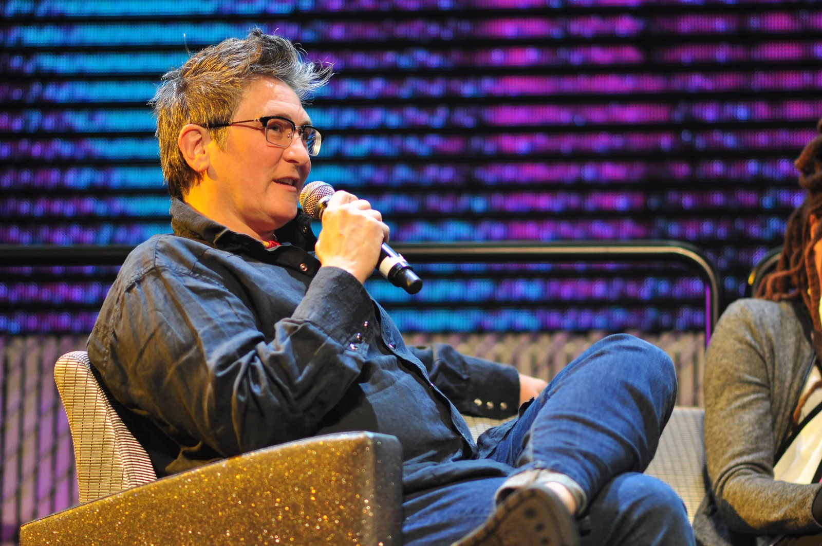 k.d. lang speaking into a microphone during the 2016 Pop Conference keynote panel in Seattle, with part of another panellist retained at right