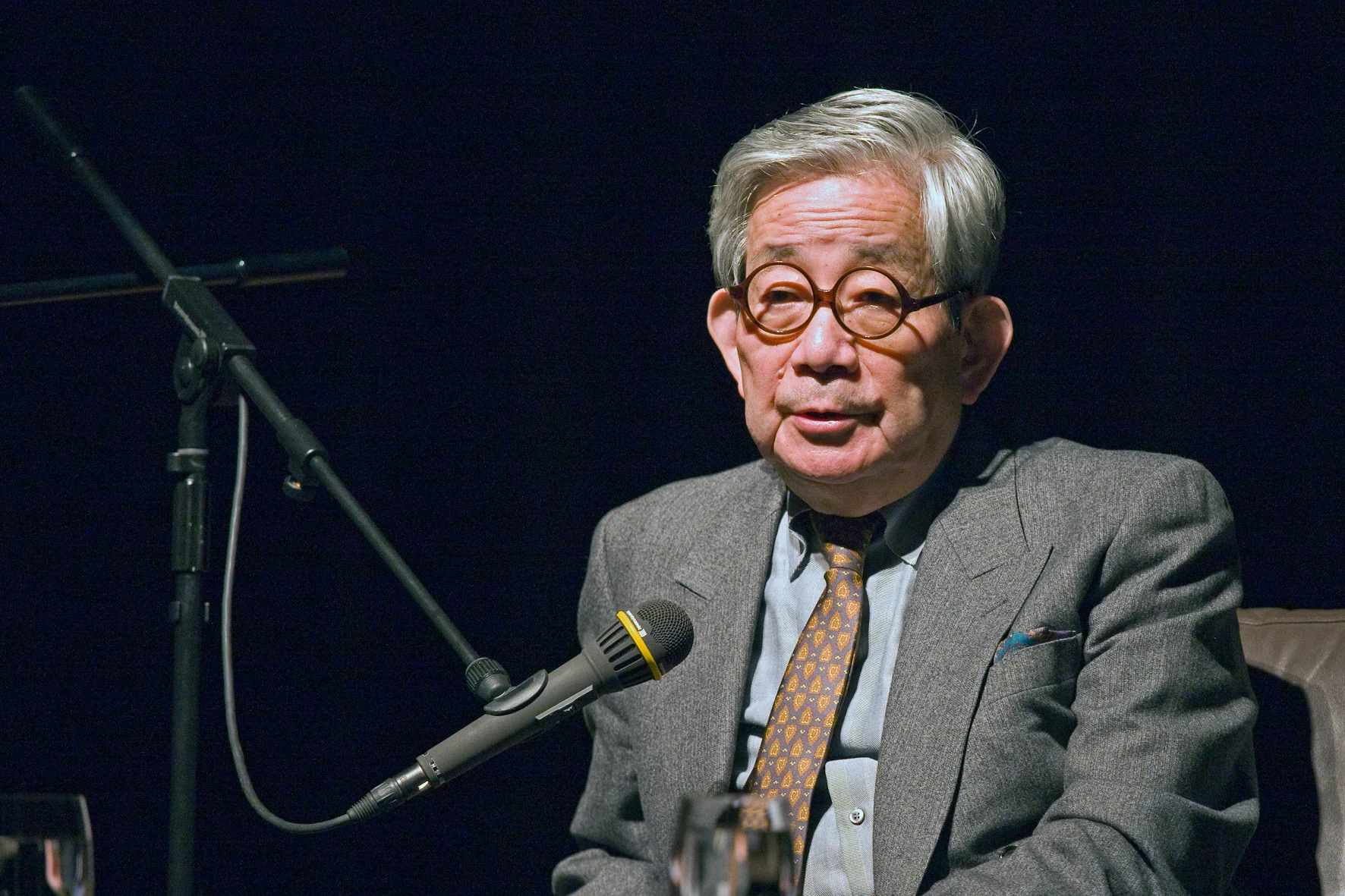 Kenzaburo Oe seated at microphones at the Japan Institute in Cologne. The full frame includes the table and dark stage around him.