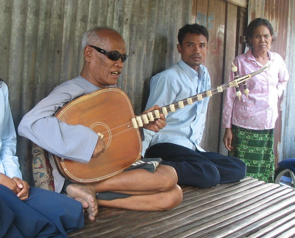 Kong Nay playing a long-necked chapei lute while seated with other people, photographed in June 2007.