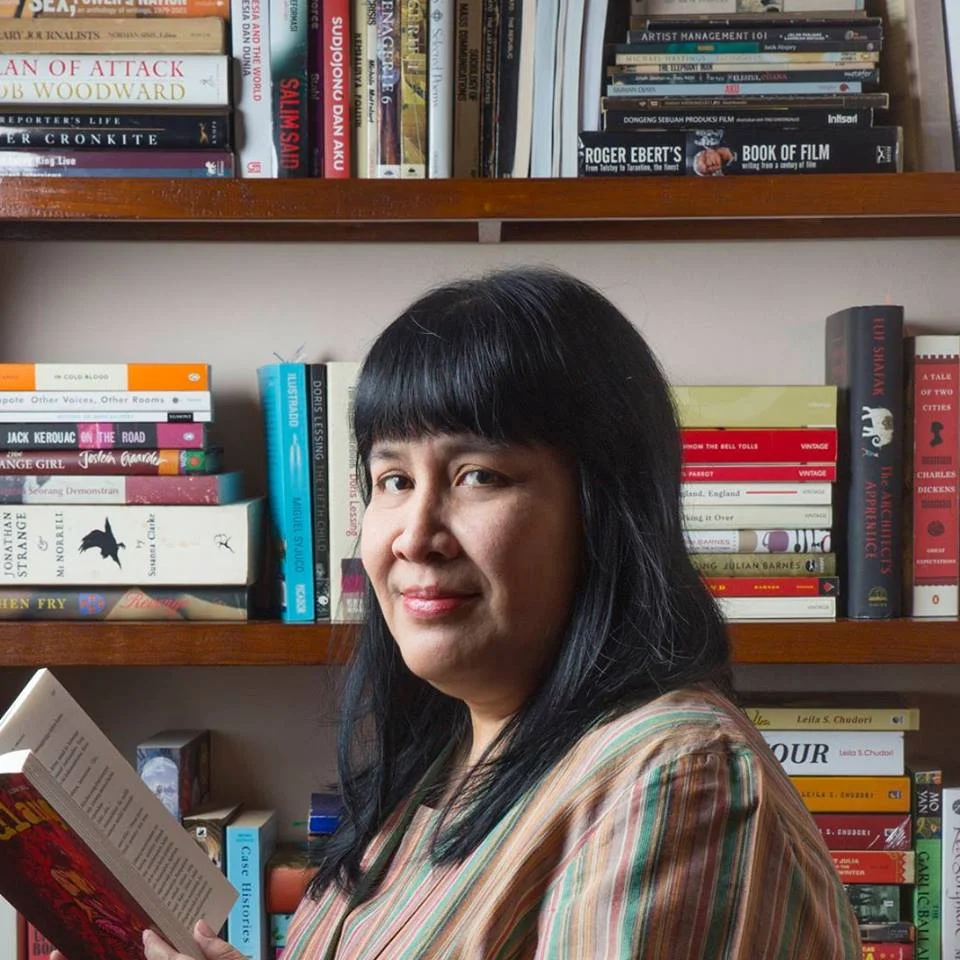 Leila Chudori seated beside bookshelves and holding a book.