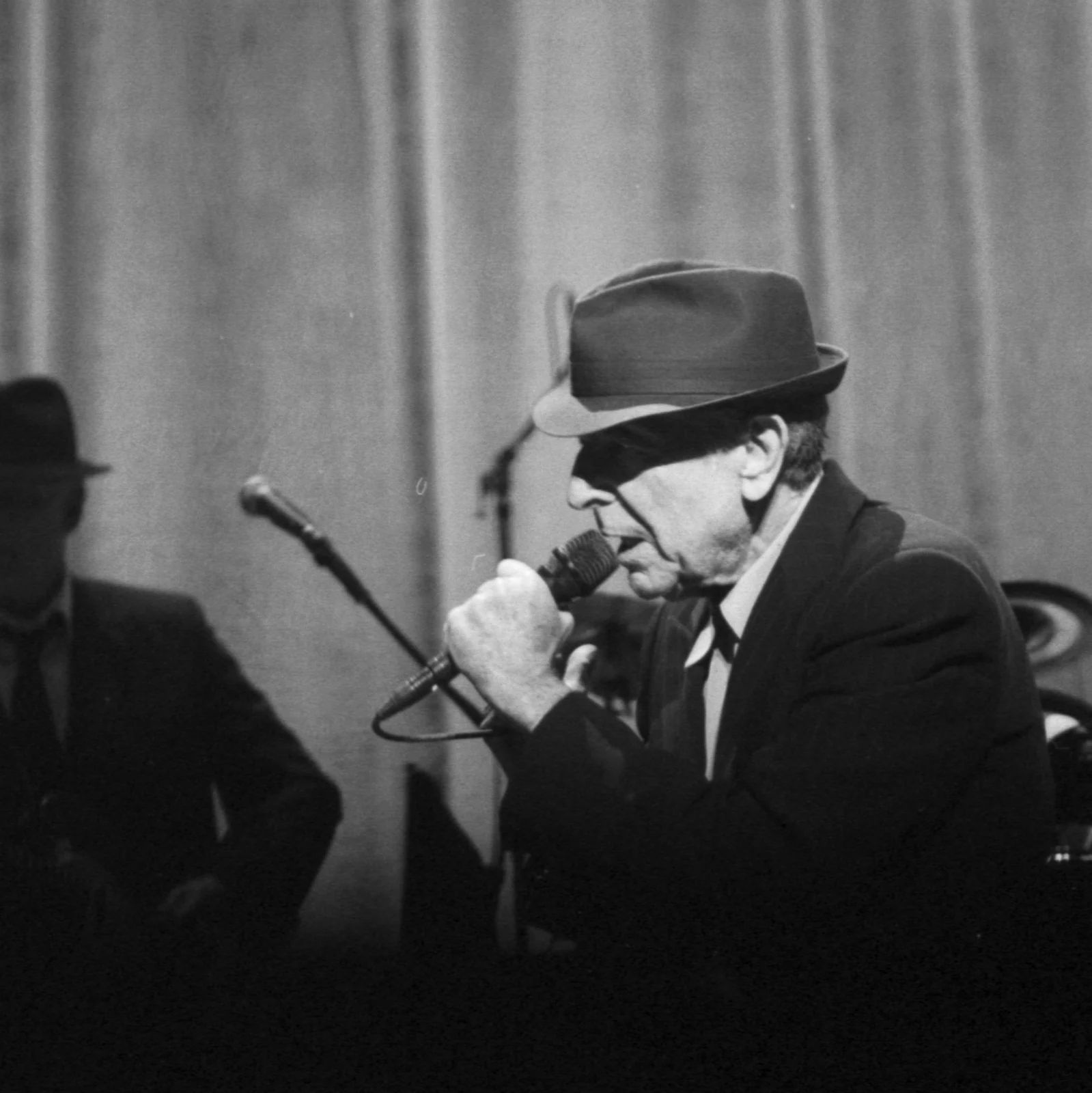 Leonard Cohen singing into a microphone during his 2008 tour, with the neighbouring musician retained in the complete frame
