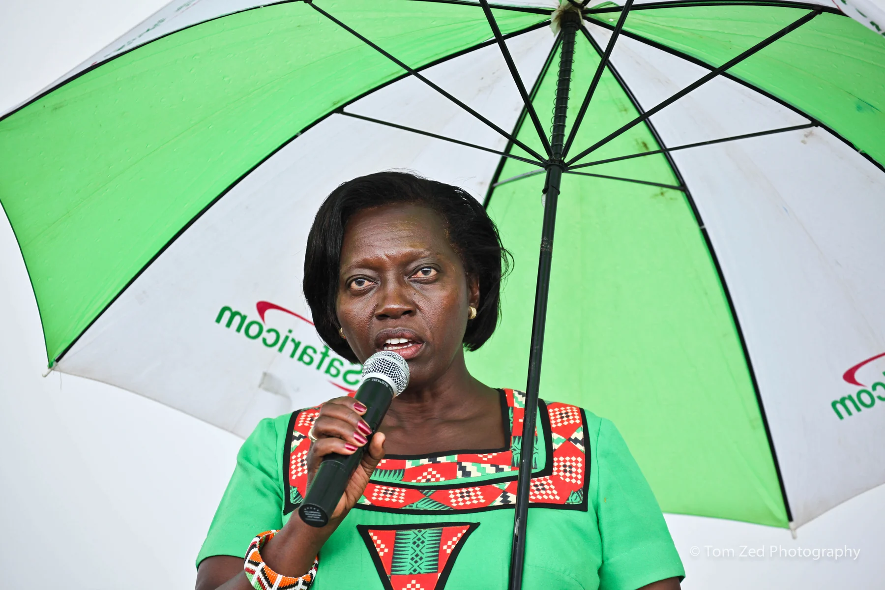 Martha Karua speaking at a microphone under an umbrella during a campaign event.