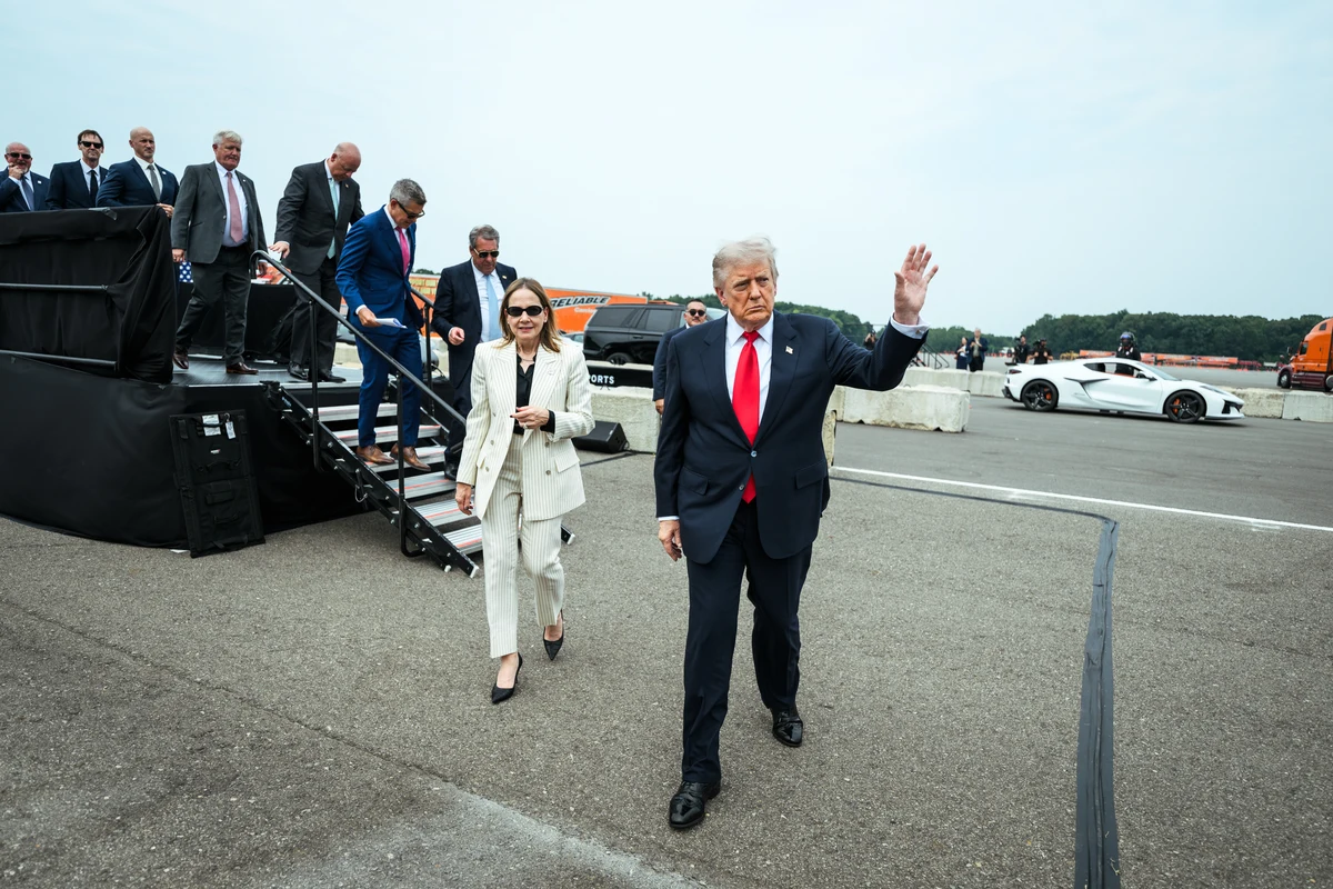 Mary Barra in a light striped suit walks left of President Donald Trump at GM's Milford proving ground on 27 July 2026, with stairs, staff and cars in the complete frame.