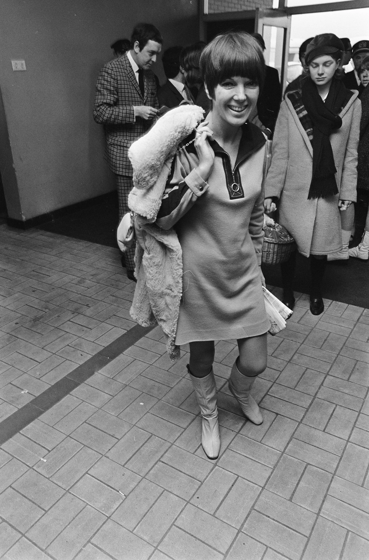 Mary Quant arriving at Schiphol in December 1966, wearing a short dress and boots with a coat and bag over her shoulder; full archival photograph.
