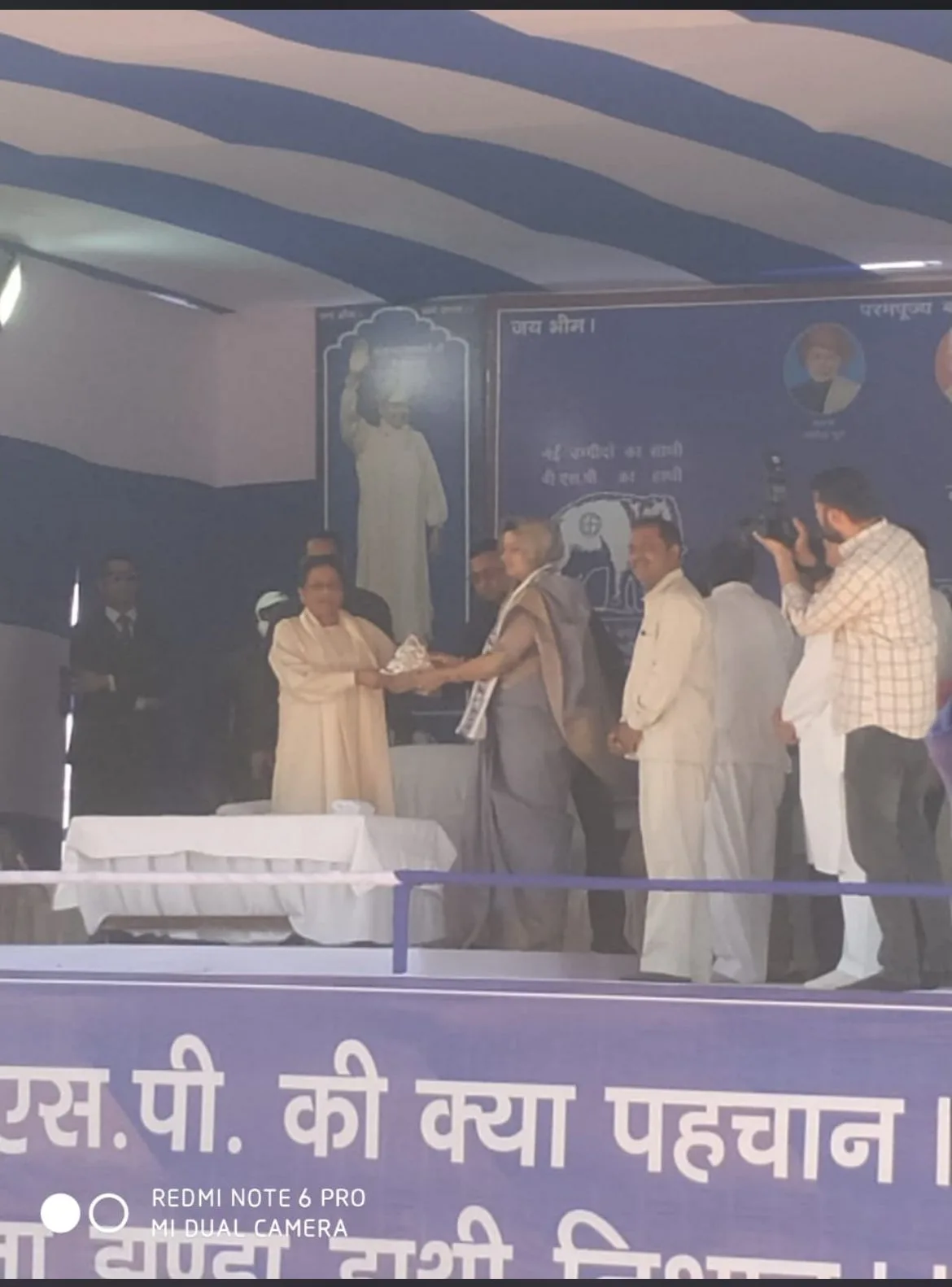 Mayawati, in cream at left of the central group, receives a presentation on an election stage; other participants and the camera watermark remain visible.