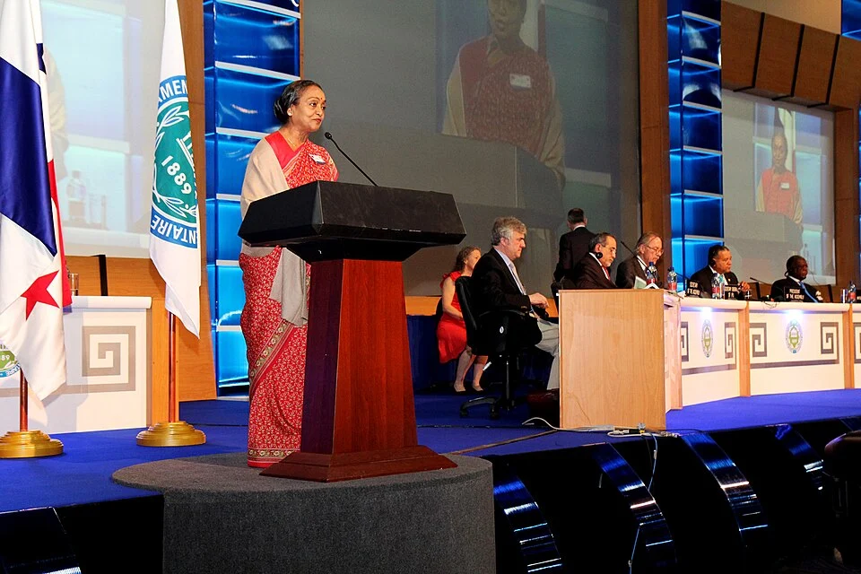 Meira Kumar stands at the left podium in a red sari at an international parliamentary assembly in Panama in April 2011; the platform and other participants remain visible.