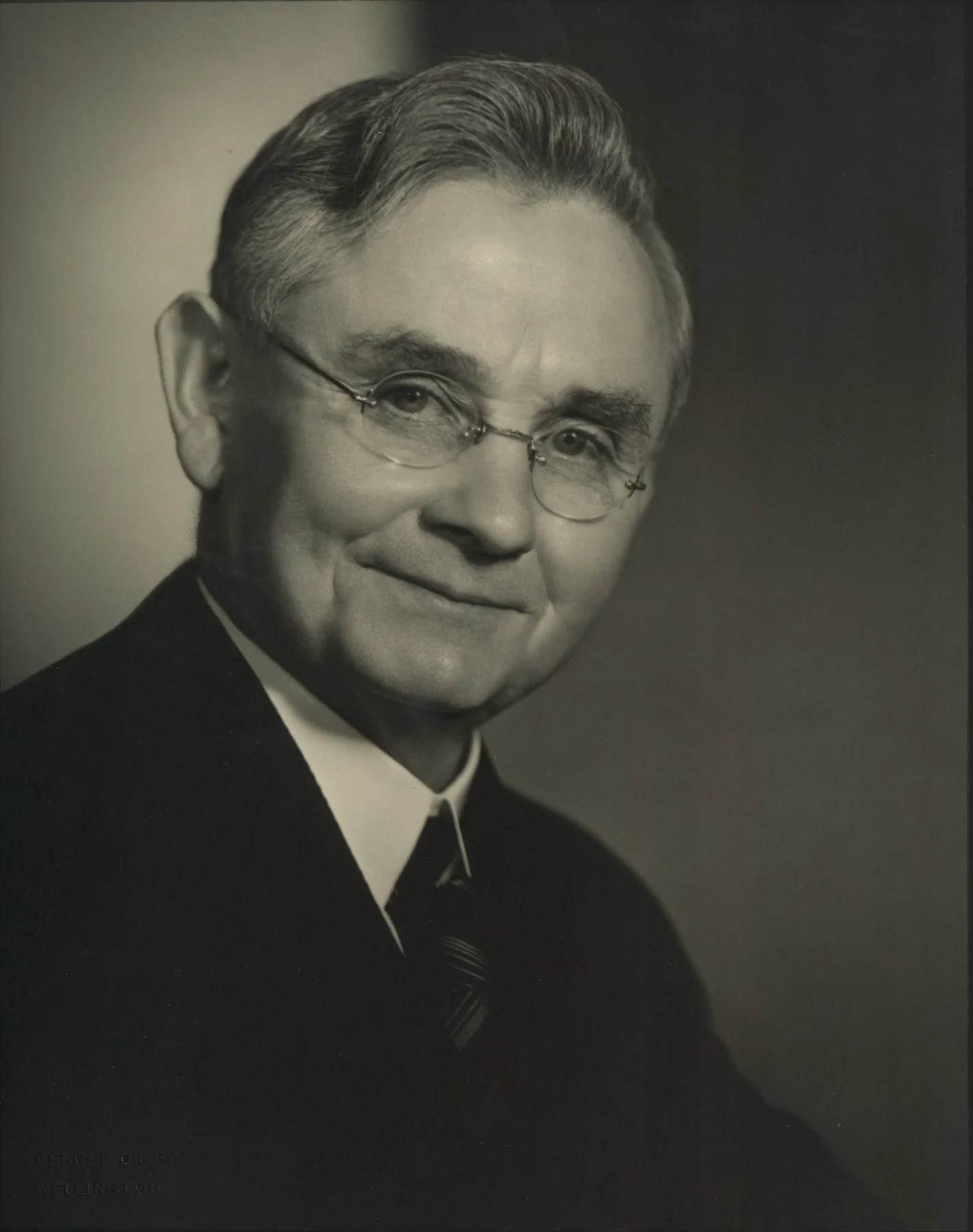 Michael Joseph Savage seated for a studio portrait, with a divided light and dark background.