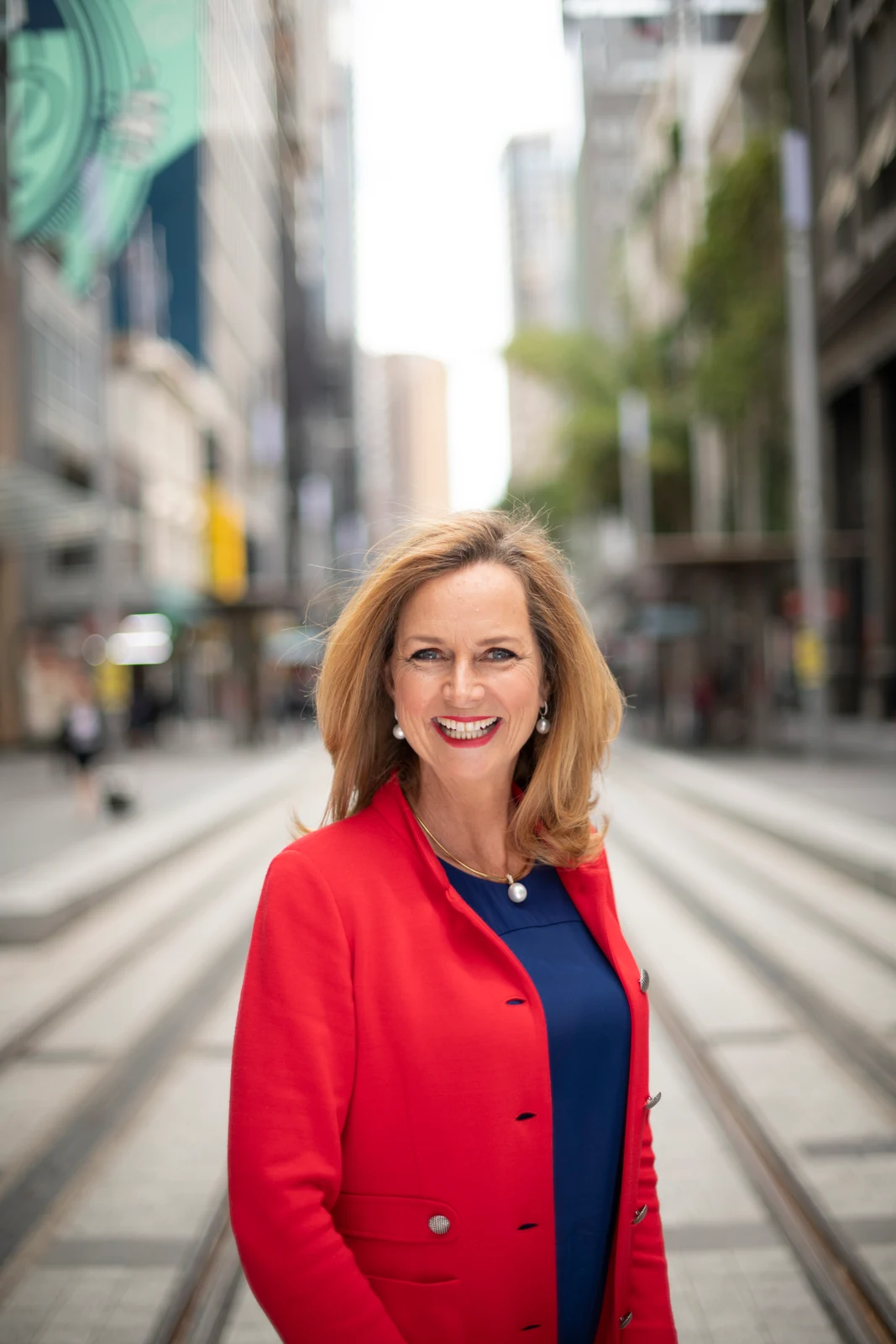Complete portrait of Naomi Simson smiling in a red jacket and navy dress beside tram tracks in Sydney CBD, with the original city background and headroom retained.