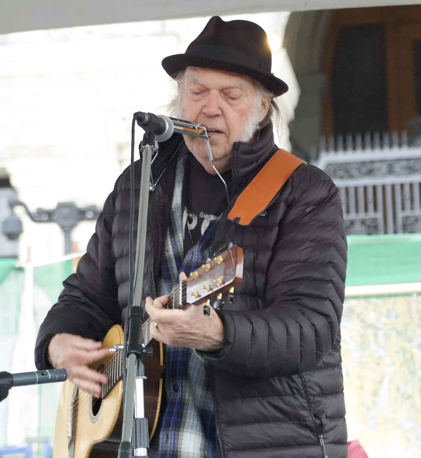 Neil Young in a dark hat and coat playing acoustic guitar and harmonica for Fairy Creek activists in 2023