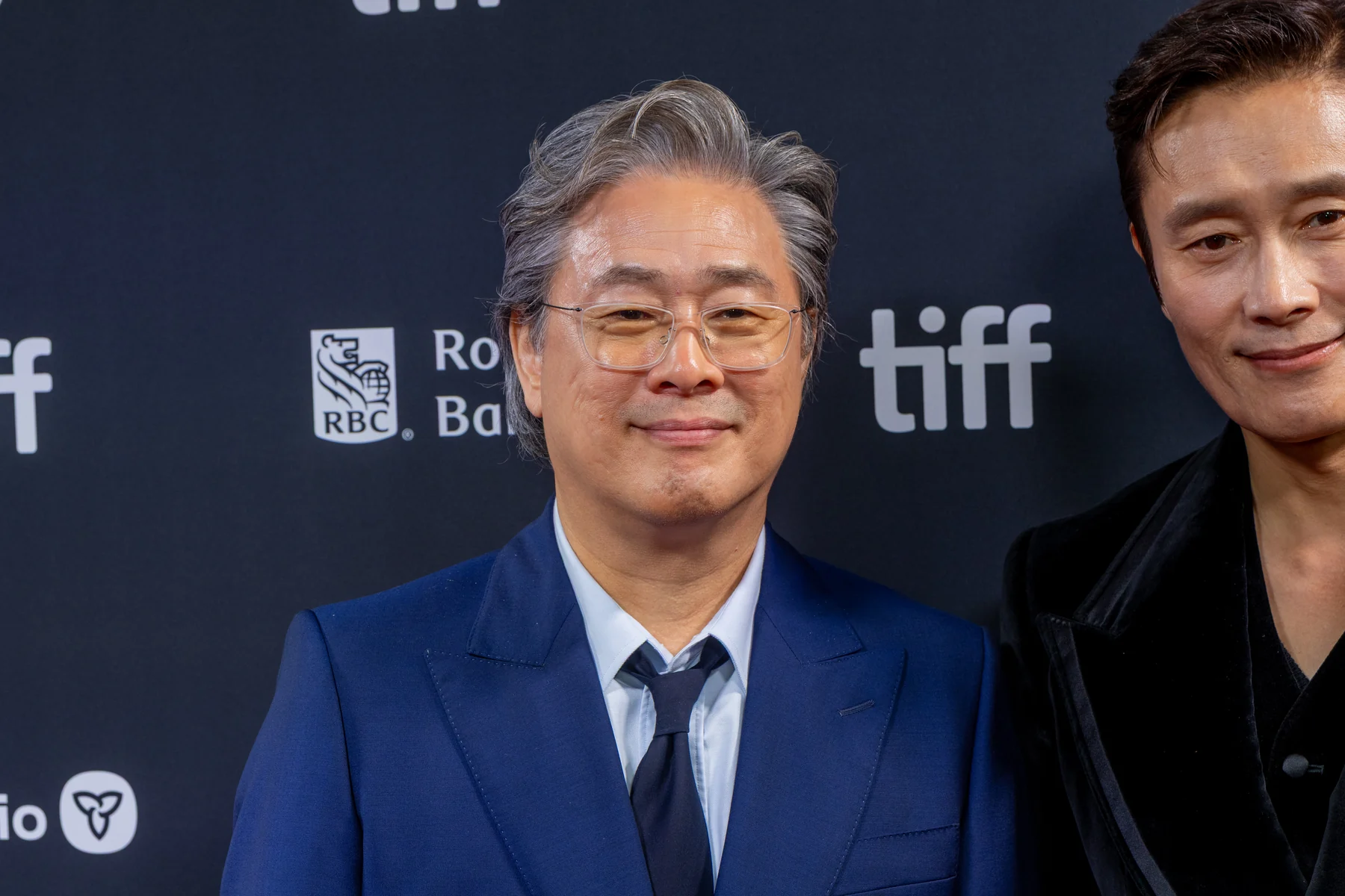 Park Chan-wook in a blue suit at the 2025 Toronto International Film Festival, with part of another participant at the right.