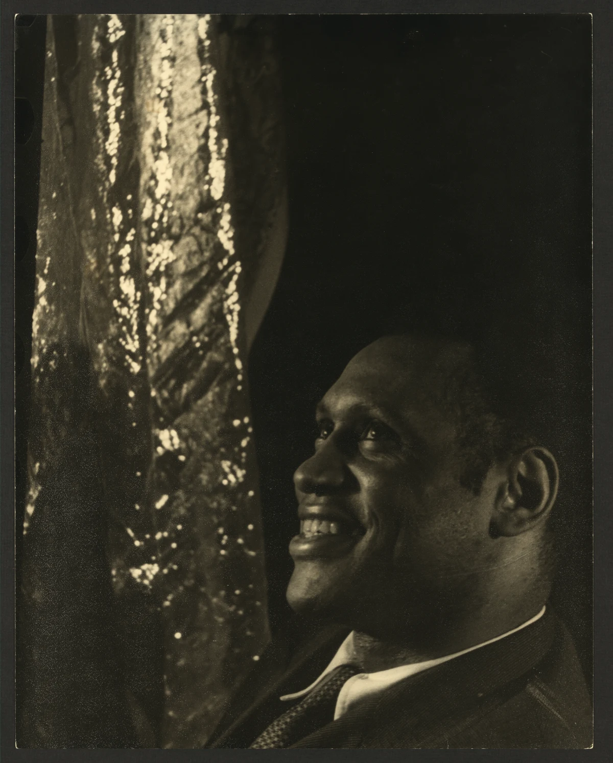 Paul Robeson smiling beside a patterned curtain in Carl Van Vechten's portrait of 1 June 1933, with the complete warm-toned print and mount edges retained.