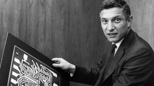 Robert Noyce in a suit holding a board with a large circuit pattern.