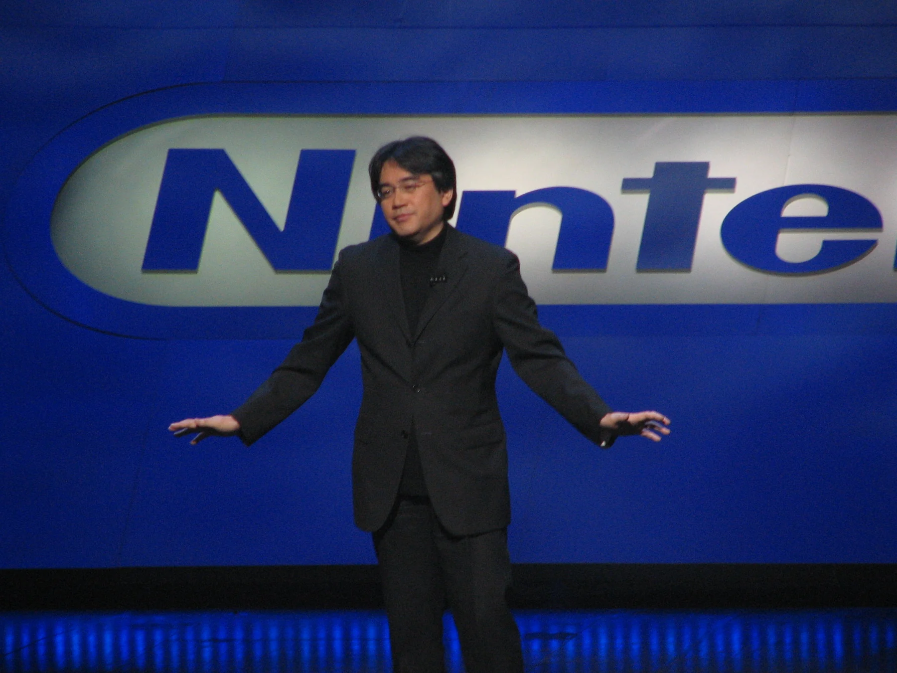 Satoru Iwata standing with his arms extended on the Nintendo E3 stage in 2006.