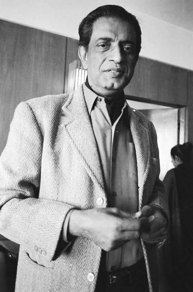 Satyajit Ray standing indoors in New York in Dinu Alam’s complete black-and-white photograph, with a second person visible in the background