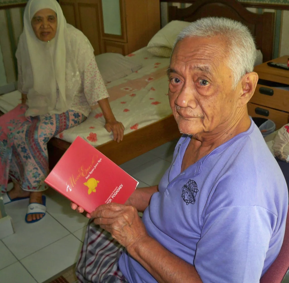 Shahnon Ahmad seated at home holding a red book, with another person visible behind him.