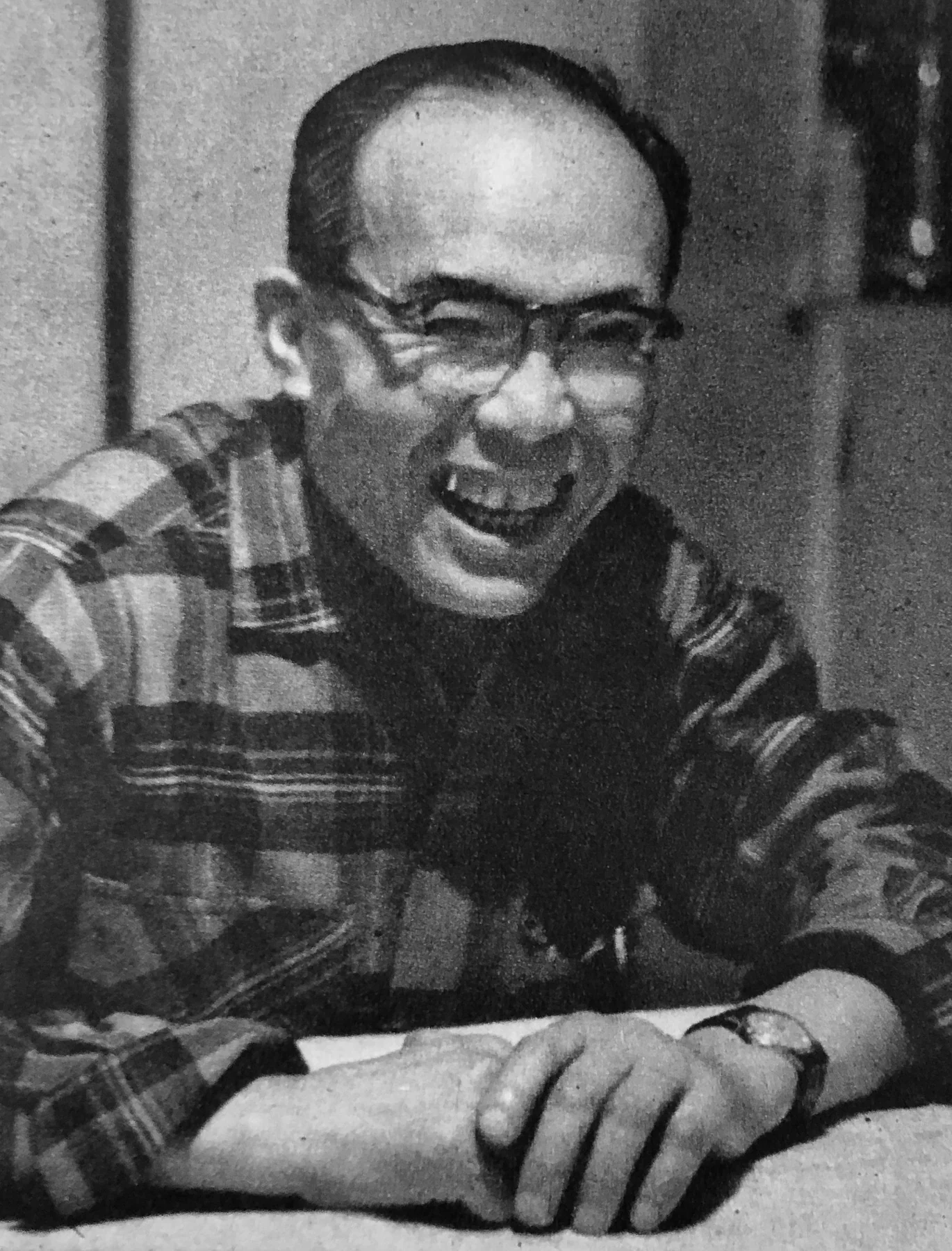 Soichiro Honda laughing while seated at a table in a checked shirt, photographed in 1954.