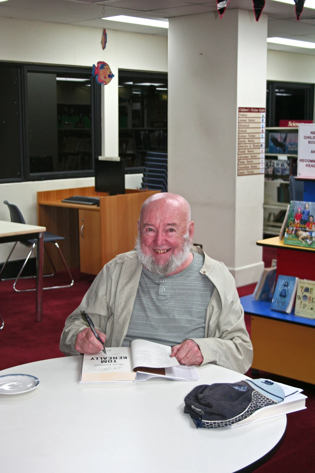 Thomas Keneally smiling while signing a book at Mosman Library in February 2011, with the full table and library surroundings retained.